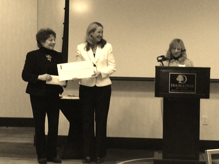 Three women stand in what appears to be a conference or seminar room. Two women on the left, one holding a large check, face the audience, while the third woman on the right speaks at a podium with a microphone. The podium has a "DoubleTree" logo.